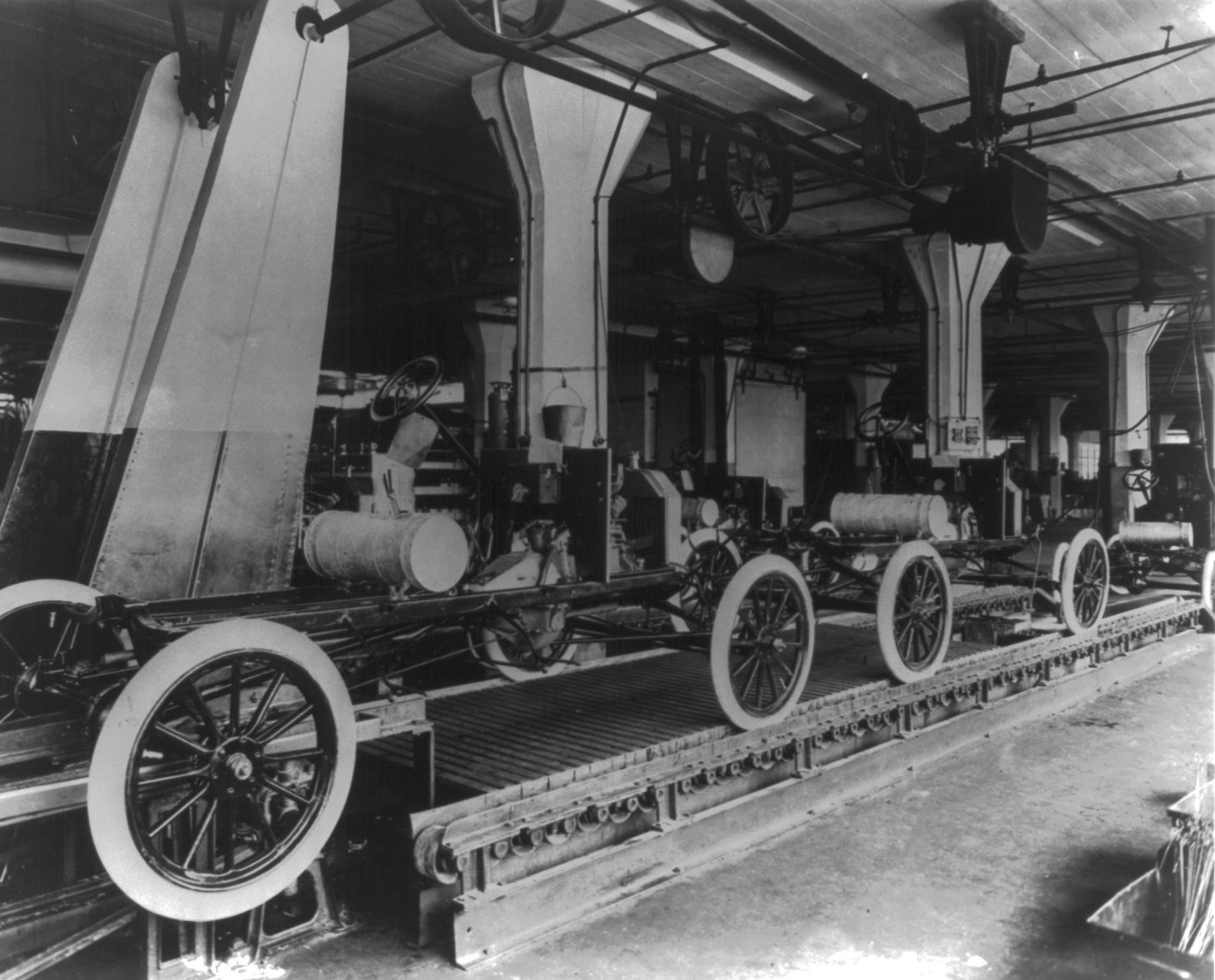 Black and white photograph of Ford Model T chassis on the moving assembly line at the Highland Park plant in 1913, with line shafting, belt pulleys and hangers running along the ceiling above